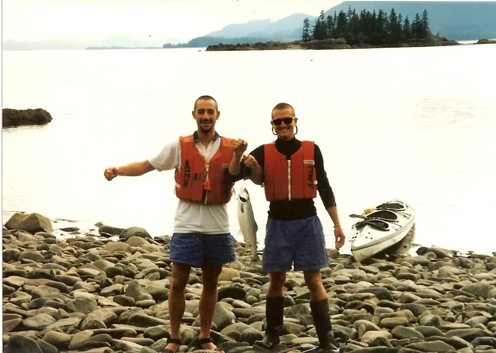 two friends in life jackets on a rocky beach, one holding up a fish, a kayak behind them
